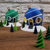 A blue and a green 3D printed Christmas bauble fairy house, lit from within, sitting on a snowy base surrounded by small green trees.
Close-up of a blue, light-up bauble-shaped house with glowing windows on a wooden table.
A whimsical 3D printed Christmas village scene with two bauble-shaped houses and small pine trees against a white brick wall.
