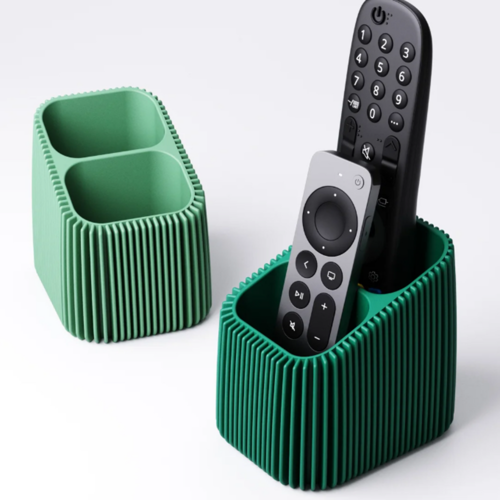 A modern green remote control holder with a vertical finned texture, holding a silver and a black TV remote on a white background.
Two 3D printed remote holders in different shades of green, showing the two-compartment design and textured finned pattern.
A dark green finned remote caddy sitting on a coffee table, neatly storing two television remotes.