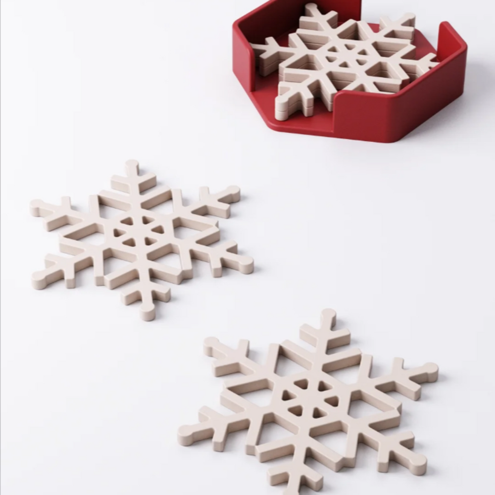 A set of four white snowflake-shaped coasters stacked neatly inside a modern red hexagonal holder on a white background.
A white snowflake coaster on a dark wood table, with a festive red mug of hot chocolate sitting on it.
A stack of white snowflake coasters next to two holder options, one red and one green.