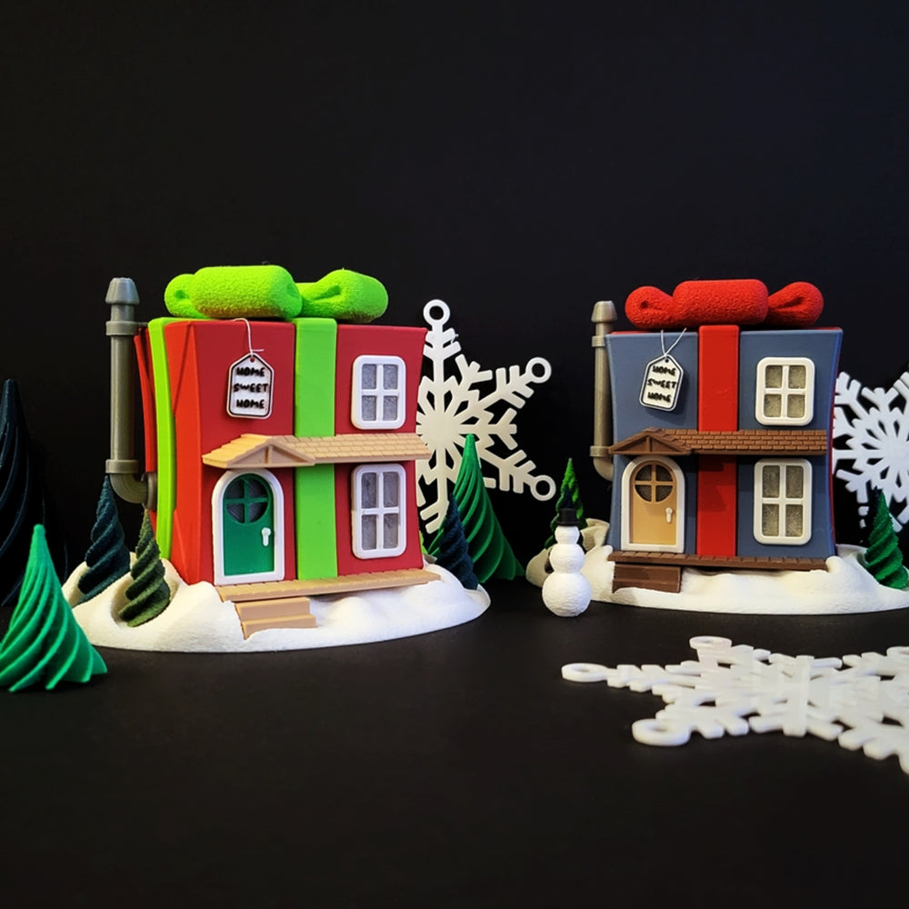 A 3D printed Christmas fairy house shaped like a red and green present with a bow, lit from within, sitting on a snowy base with small trees.
A blue and a red light-up gift box house, shown in a festive scene with a snowman and small trees on a dark background.
Close-up of the "Home Sweet Home" sign hanging on the door of a red, present-shaped fairy house.