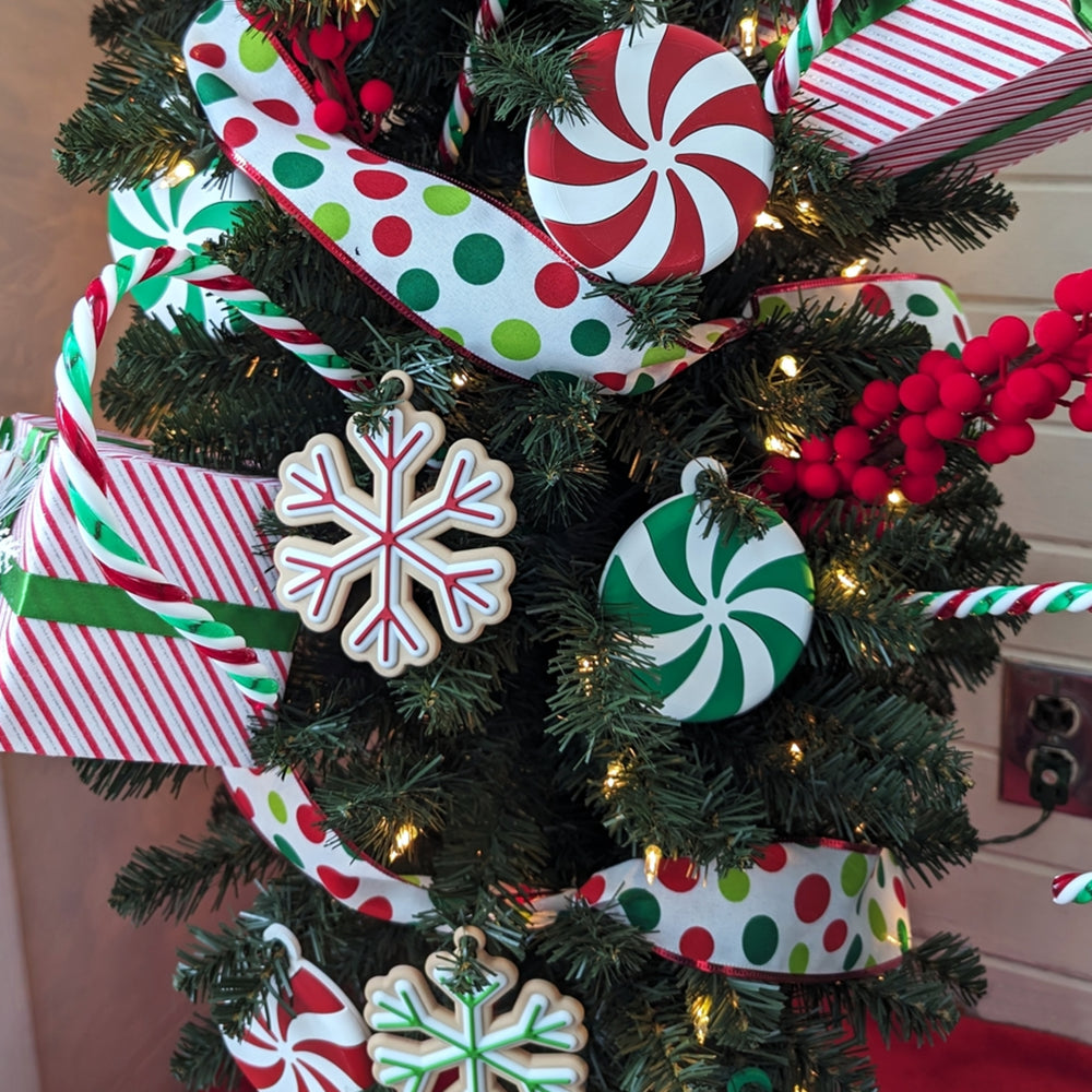 A 3D printed peppermint swirl ornament with red and white sections, lying on a dark wood grain surface. A set of three red and white candy-style Christmas tree ornaments with hanging loops. Close-up showing the 3D printed texture of a red and white peppermint ornament.