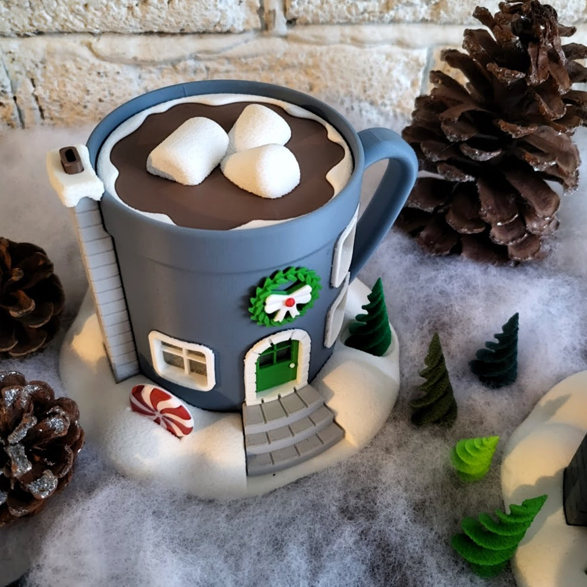 A 3D printed Christmas fairy house shaped like a blue mug of hot chocolate, with glowing windows, a wreath, and marshmallow roof, set in a snowy scene.
A whimsical light-up hot cocoa mug house, shown on a snowy base with small trees and pinecones against a white brick wall.
Close-up of the festive details on a hot chocolate mug house, including a small green door, wreath, and candy cane.