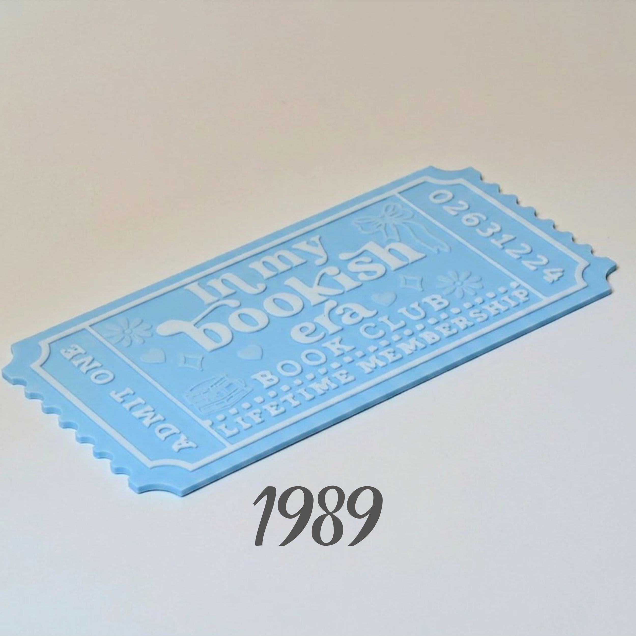 A light blue ticket-shaped book club membership card with embossed text reading 'In my bookish era BOOK CLUB LIFETIME MEMBERSHIP' and the number '02631224', shown on a plain light background with '1989' printed below.