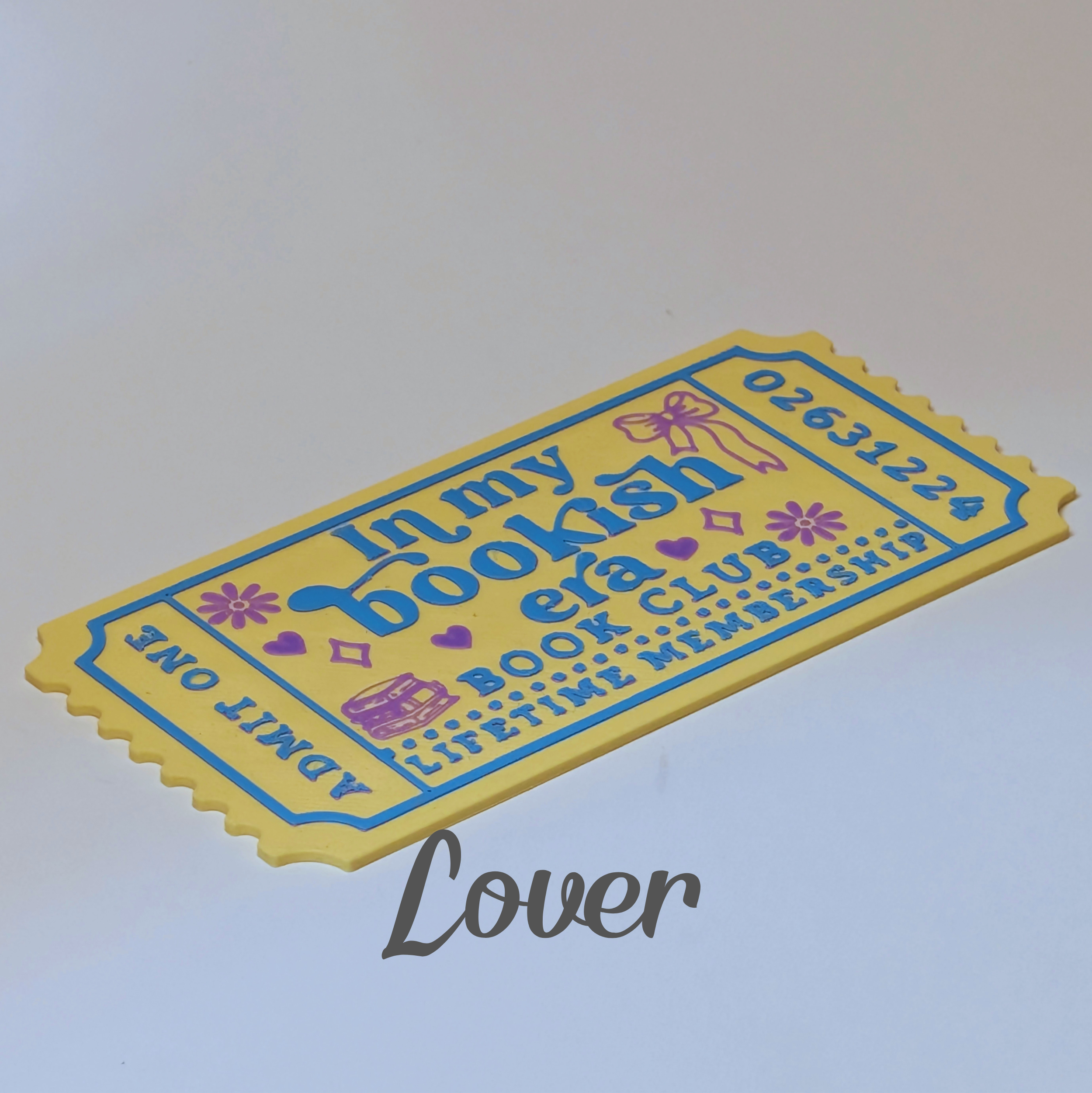 A yellow and blue bookish ticket-shaped mat or plaque with decorative text reading 'In my bookish era book club lifetime membership' and 'Admit one', shown on a plain light background with a 'Lover' watermark.