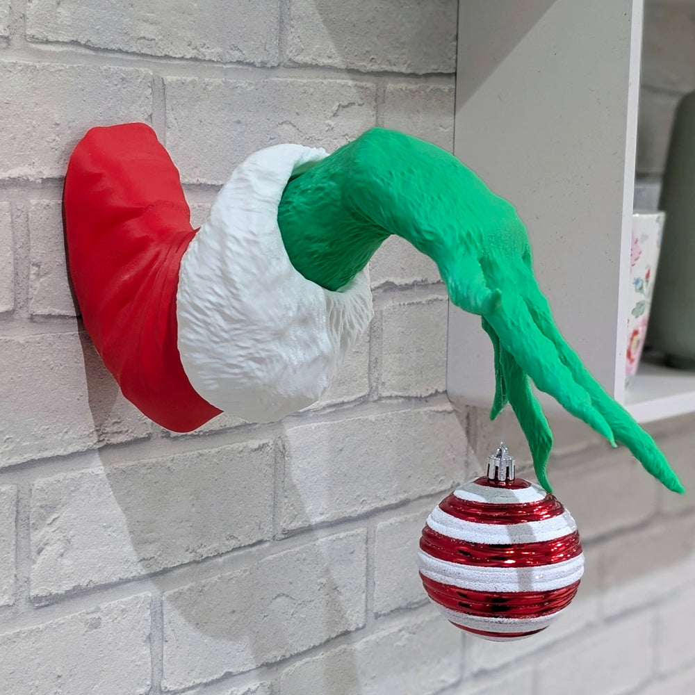 A green grinch hand in a red and white santa cuff, appearing to come out of a white brick wall while holding a red and white striped Christmas bauble.
Side view of a 3D printed green hand wall decoration, showing its depth and how it holds a Christmas ornament.
A fun and quirky Christmas wall decoration shaped like a mischievous green hand reaching out from the wall.