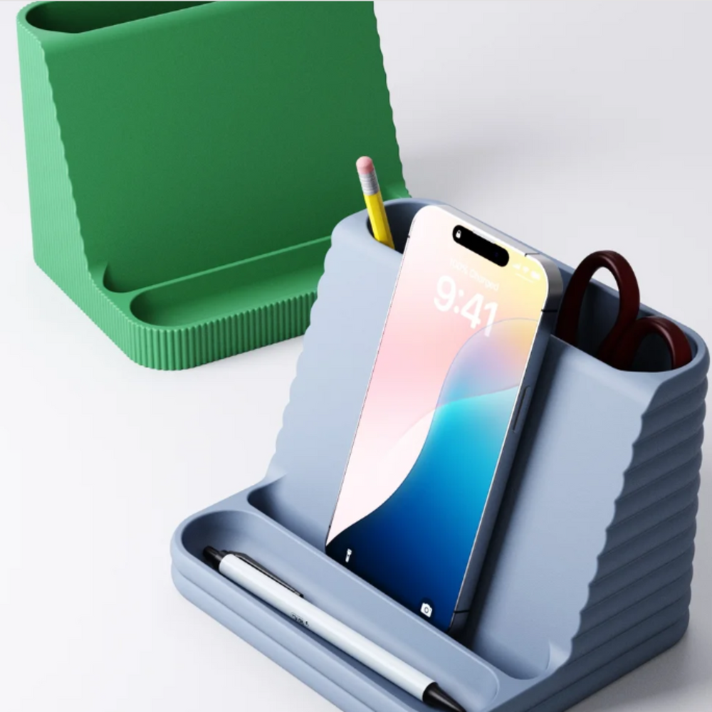 A blue desk organiser with horizontal flutes, holding a smartphone, pens, and scissors on a white background.
A green desk organiser with vertical ribs in the background, shown next to the blue version.
Lifestyle photo of the desk tidy on a modern white desk, keeping a phone and stationery organised next to a laptop.