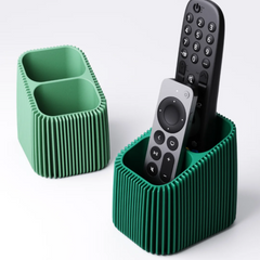 A modern green remote control holder with a vertical finned texture, holding a silver and a black TV remote on a white background.
Two 3D printed remote holders in different shades of green, showing the two-compartment design and textured finned pattern.
A dark green finned remote caddy sitting on a coffee table, neatly storing two television remotes.