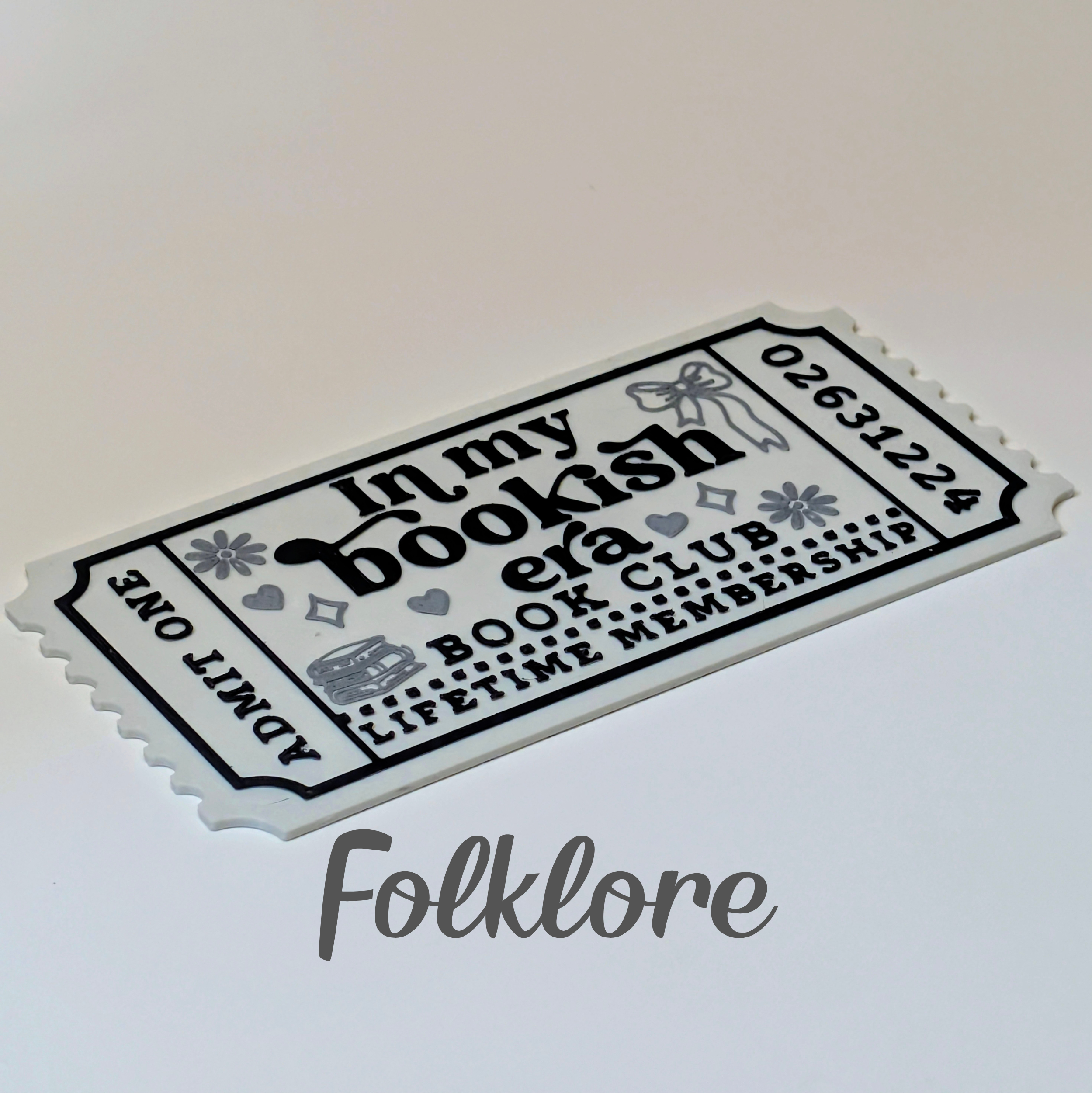 A white paper-style ticket with black decorative text reading 'In my bookish era BOOK CLUB LIFETIME MEMBERSHIP' and 'ADMIT ONE,' photographed on a light neutral background. A gray 'Folklore' watermark appears below the ticket.